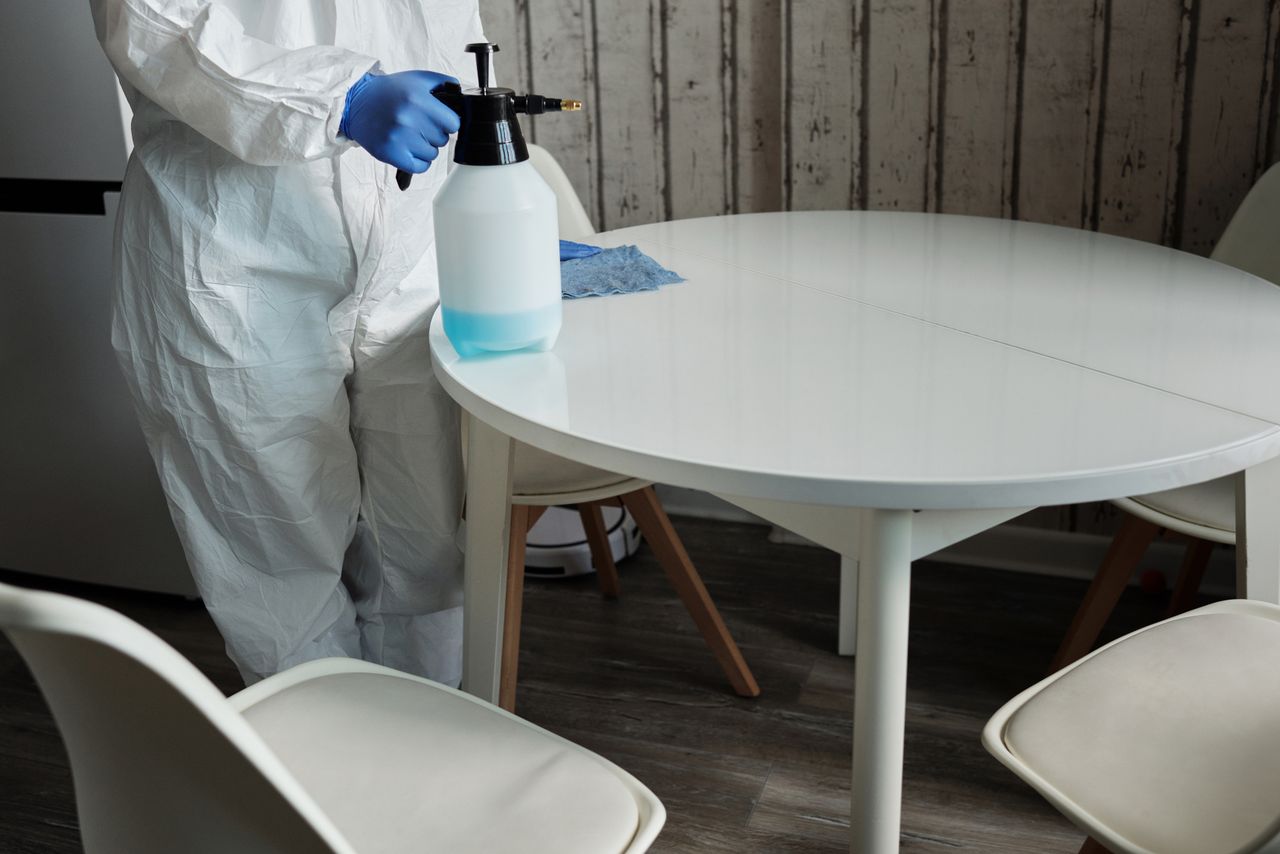why-choose-us Person in protective suit disinfecting a white table indoors with a sprayer.